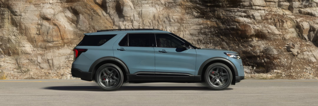 A sleek white 2025 Ford Explorer driving on a scenic mountain road, showcasing its dynamic design and premium features. The SUV features bold mesh grille, signature lighting, and 21-inch magnetite painted wheels, highlighting its sophistication and flair.